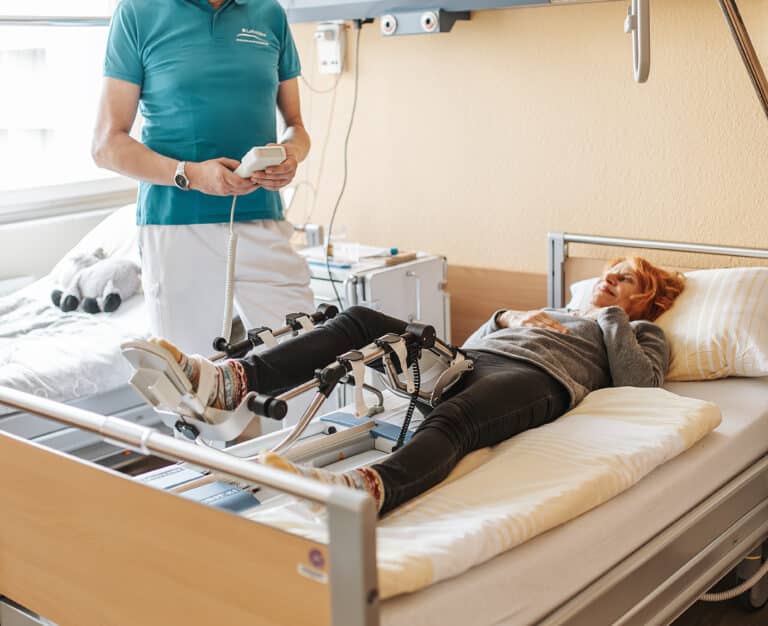 887A1308-Bearbeitet Ein Arzt überprüft die Mobilität des rechten Beines einer Patientin mit Hilfe eines Geräts, welches das Bein auf Knopfdruck höher lagern kann.